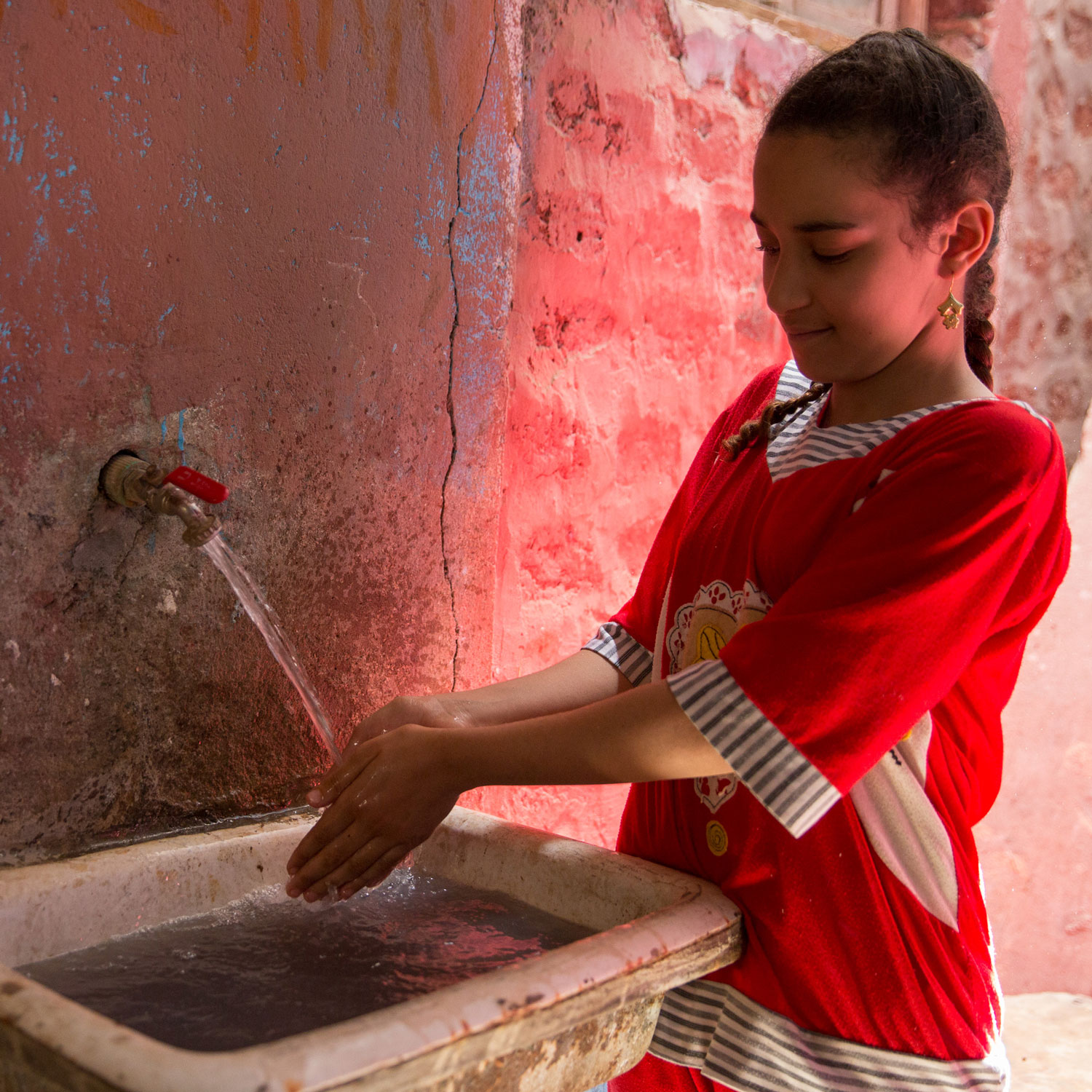  Esraa, 11, washes her hands in her home in Beni Edrees, while Heba talks to her mother about her satisfaction with the waste water services.  She shared they now shower much more often as they no longer have to worry about their septic tank overflowing. Their tank used to fill up every week, which was very expensive to drain.   Photo for USAID/Egypt.  